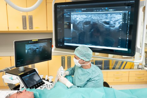 Patient liegt wach mit OP-Tuch bedeckt am Tisch, Arzt führt mit Blick auf Bildschirm Eingriff durch
