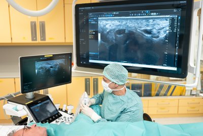 Patient liegt wach mit OP-Tuch bedeckt am Tisch, Arzt führt mit Blick auf Bildschirm Eingriff durch