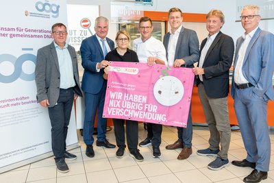 Sechs Männer und eine Frau stehen nebeneinander und halten ein rosa Transparten mit der Aufschrift "Wir haben nix übrig für Verschwendung".