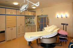 Gebärzimmer mit einem beweglichen Gebärbett, mit hellgelber Bettwäsche, Wandlampen, große Deckenlampe, Kastenverbau, Waschbecken.