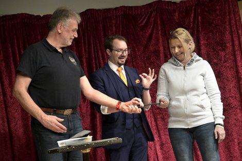 Zauberkünstler Harald Brunner („Harry Fontaine“) führt auf einer kleinen Bühne vor rotem Vorhang einen Trick mit Ring und Stab vor – zwei Personen stehen neben ihm und beobachten die Vorführung aus nächster Nähe.