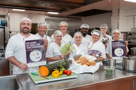 Gruppenfoto Küchenteam vor der Arbeitsplatte mit Lebensmittel und Broschüre von United Against Waste