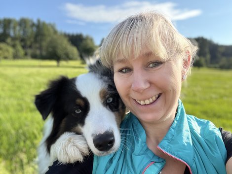 Selfie von Sylvia Hollnsteiner in der Natur mit ihrem Hund, der seien Kopf liebevoll auf ihre Schulter legt