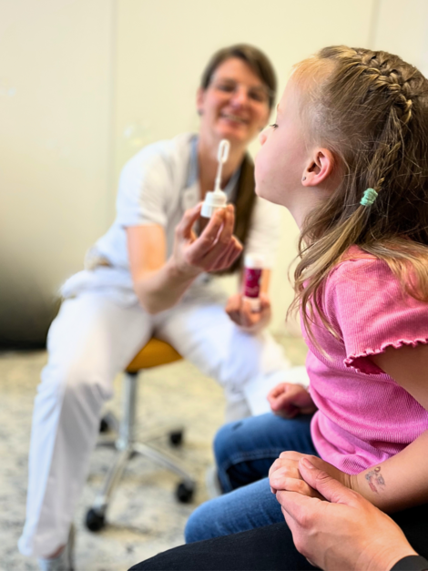 Krankenpflegerin hält Seifenblasen-Stäbchen, kleines Mädchen sitzt am Schoss der Mama und bläst durch