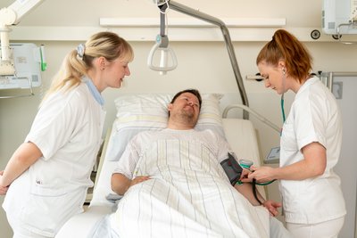 Pflegerin berät Auszubildende beim Blutdruckmessen bei einem Patienten