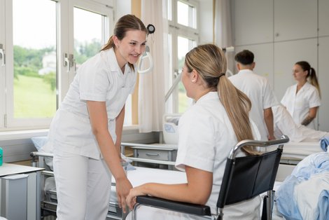 2 Pflegeschülerinnen, eine davon sitzt im Rollstuhl, im Hintergrund Krankenzimmer und weitere PflegeschülerInnen