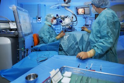 Im Operationssaal führen zwei Personen in steriler Schutzkleidung eine Augenoperation durch. Eine Person arbeitet an einem Operationsmikroskop, während die zweite Person assistiert. Moderne medizinische Geräte und Monitore umgeben den Operationsbereich.