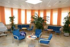Wintergarten mit blauen Stühlen, großen grünen Zimmerpflanzen, kleinen Tischen und einem Bett. Auf den Tischen sind Zeitschriften. Vorhänge sind orange.