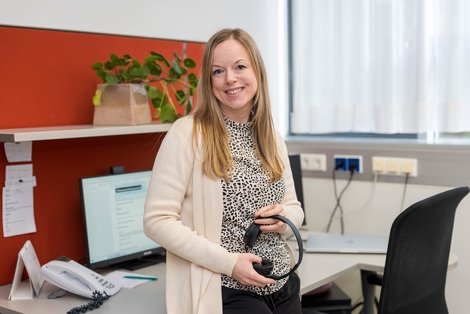 Andrea Nußbaumer steht an einem Büroarbeitsplatz, hält Kopfhörer in der Hand und blickt zur Kamera; im Hintergrund sind Computer, Telefon und eine Pflanze auf einem Regal zu sehen.