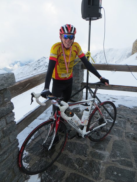Dominik Schickmair steht in Radbekleidung mit einer Medaille um den Hals mit seinem Rennrad auf einer Aussichtsplattform in einer verschneiten Berglandschaft.