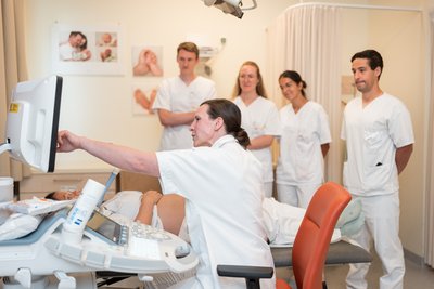 Medizinstudenten stehen um Liege, auf der Schwangere liegt. Im Vordergrund erklärt Ärztin am Bildschirm die Ultraschallbilder