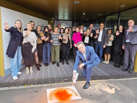 Geschäftsführer sprayt orange Hände auf Gehtsteig, dahinter stehen MitarbeiterInnen mit ausgestreckter Hand als Stop-Zeichen gegen Gewalt
