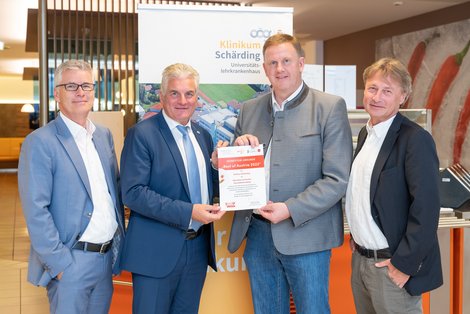 Vier Männer stehen nebeneinander. Die beiden mittleren halten eine Urkunde mit ihren Händen.