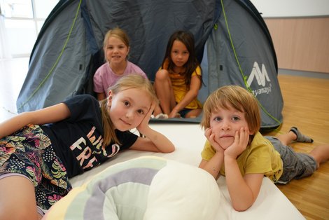 Kinder entspannen und spielen gemeinsam im Innenbereich der Sommerkinderbetreuung im Salzkammergut Klinikum Bad Ischl vor einem aufgebauten Spielzelt.
