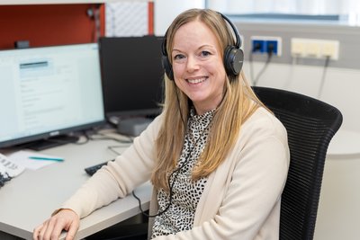 Andrea Nußbaumer sitzt an einem Schreibtisch im Büro, trägt Kopfhörer und arbeitet am Computer; Telefon, Monitor und Notizen sind sichtbar.