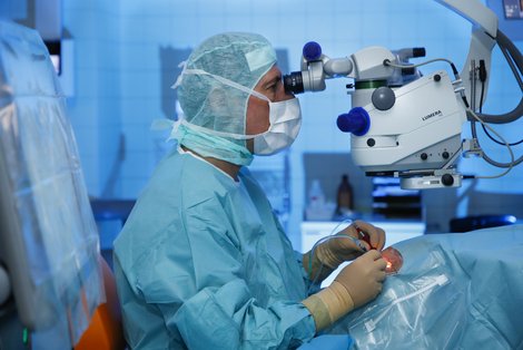 Nahaufnahme einer Person in steriler Schutzkleidung, die durch ein Operationsmikroskop blickt und eine Augenoperation durchführt. Die Hände halten chirurgische Instrumente, während der Eingriffsbereich mit sterilen Tüchern abgedeckt ist.