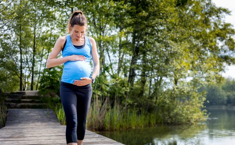 Schwangere in Sportbekleidung geht auf einem Steg am See und hält ihren Bauch