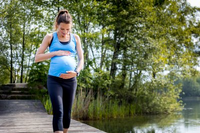 Schwangere in Sportbekleidung geht auf einem Steg am See und hält ihren Bauch