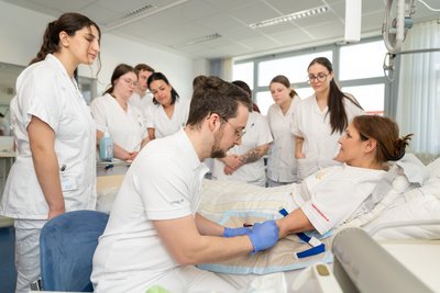 Auszubildende stehen rund um Krankenbett und üben Blutabnahme