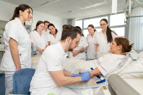 Auszubildende stehen rund um Krankenbett und üben Blutabnahme