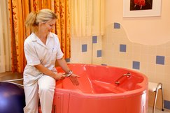 Hebamme sitzt am Rand der rosa Gebärwanne und lässt Wasser mit dem Duschkopf ein. Bild an der Wand mit rosa Blumen, Orangefarbene Vorhänge. Fliesen an der Wand in blau weiß.