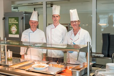 Drei Köche in Kochjacke und mit Kochmütze hinter der Essensausgabe