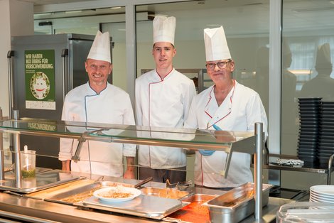 Drei Köche in Kochjacke und mit Kochmütze hinter der Essensausgabe