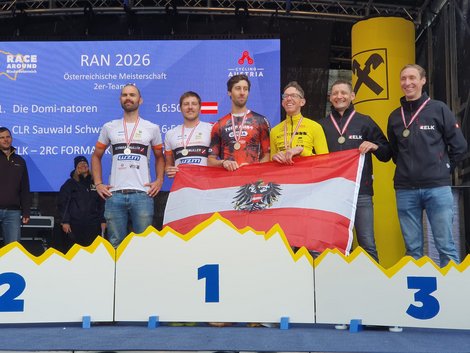 Siegerehrung eines Ultra-Radrennens: Mehrere Athleten stehen mit Medaillen auf dem Podest und halten eine österreichische Flagge. Im Hintergrund informiert eine Anzeigetafel über die Platzierungen beim Race Around Niederösterreich.