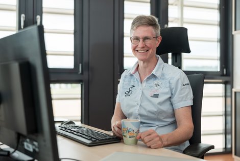 Ruth Bahmann sitzt in der MIT an ihrem Schreibtisch und sieht lächelnd in die Kamera