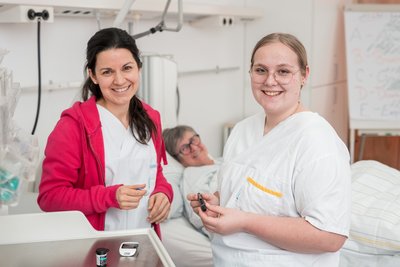zwei Auszubildende, Krankenbett im Hintergrund