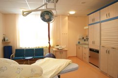 Gebärzimmer mit einem Gebärbett, mit hellgelber Bettwäsche, Babywaage, Radio, Tuch von der Decke hängend, Waschplatz, 3er Sesselreihe blau.