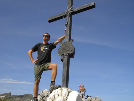 Michael Kerschhofer auf einer Bergwanderung neben einem Gipfelkreuz in alpiner Landschaft.