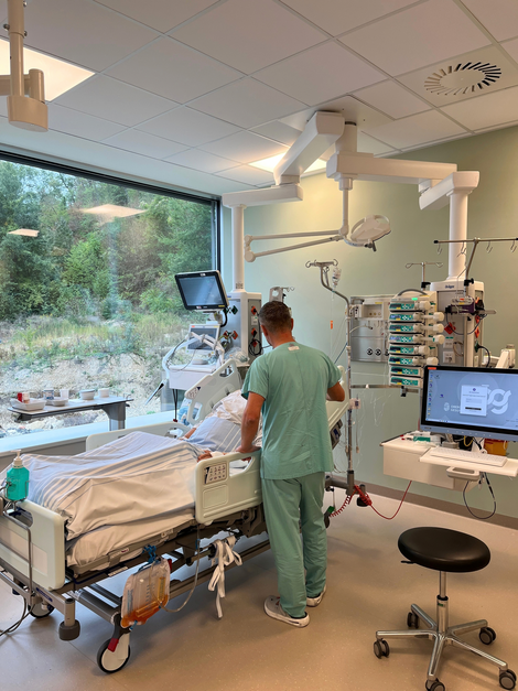Blick auf Patientenbett, Pfleger steht davon und dahinter großes Panoramafenster