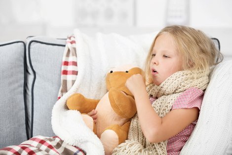 Mädchen liegt in Decke eingewickelt mit Teddy im Arm auf der Couch