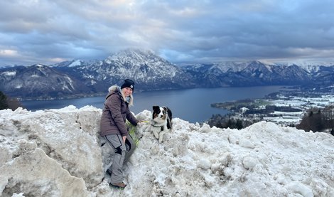 Sylvia Hollnsteiner mit ihrem Hund im Winter auf dem Gipfel eines Bergers. Im Hintergrund ein See und Berge