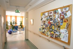 Gang mit Handlauf holzfarben an der Wand. Pinnwand mit vielen Grußkarten mit Babyfotos, Sicht auf den Wintergarten mit blauen Stühlen, Pflanzen, Peziball in lila, kleiner Tisch mit Infomaterial.