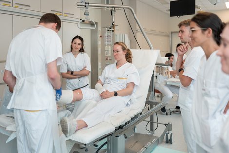 Studenten stehen um Liege, auf der Studentin ein Bein eingegipst bekommt