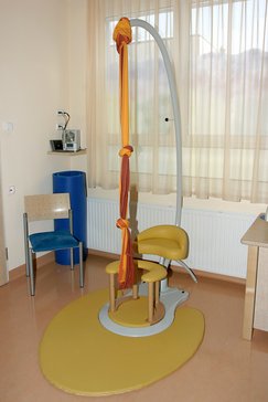 Gebärstuhl gelb. Orange/Rotes Tuch von der Decke hängend. Blauer Stuhl. Radio. Blaue weiche Matte.
