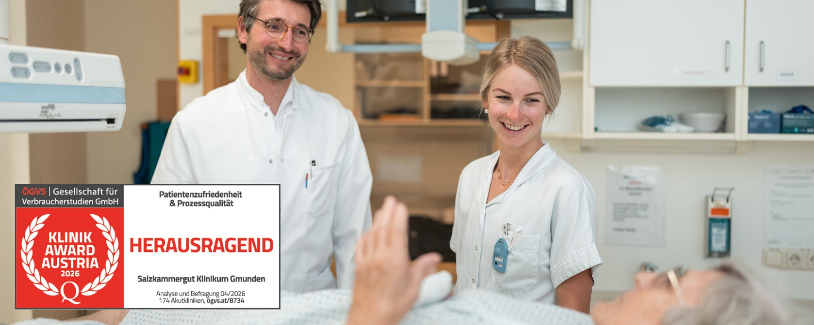Zwei Personen aus dem medizinischen Team sprechen mit einer liegenden Patientin in einem Untersuchungszimmer; sichtbar ist die Auszeichnung Klinik Award Austria – Herausragend