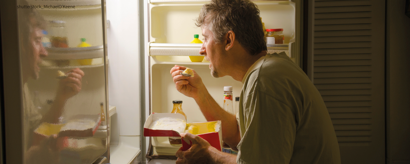 Mann sitzt vor dem Kühlschrank und isst etwas.