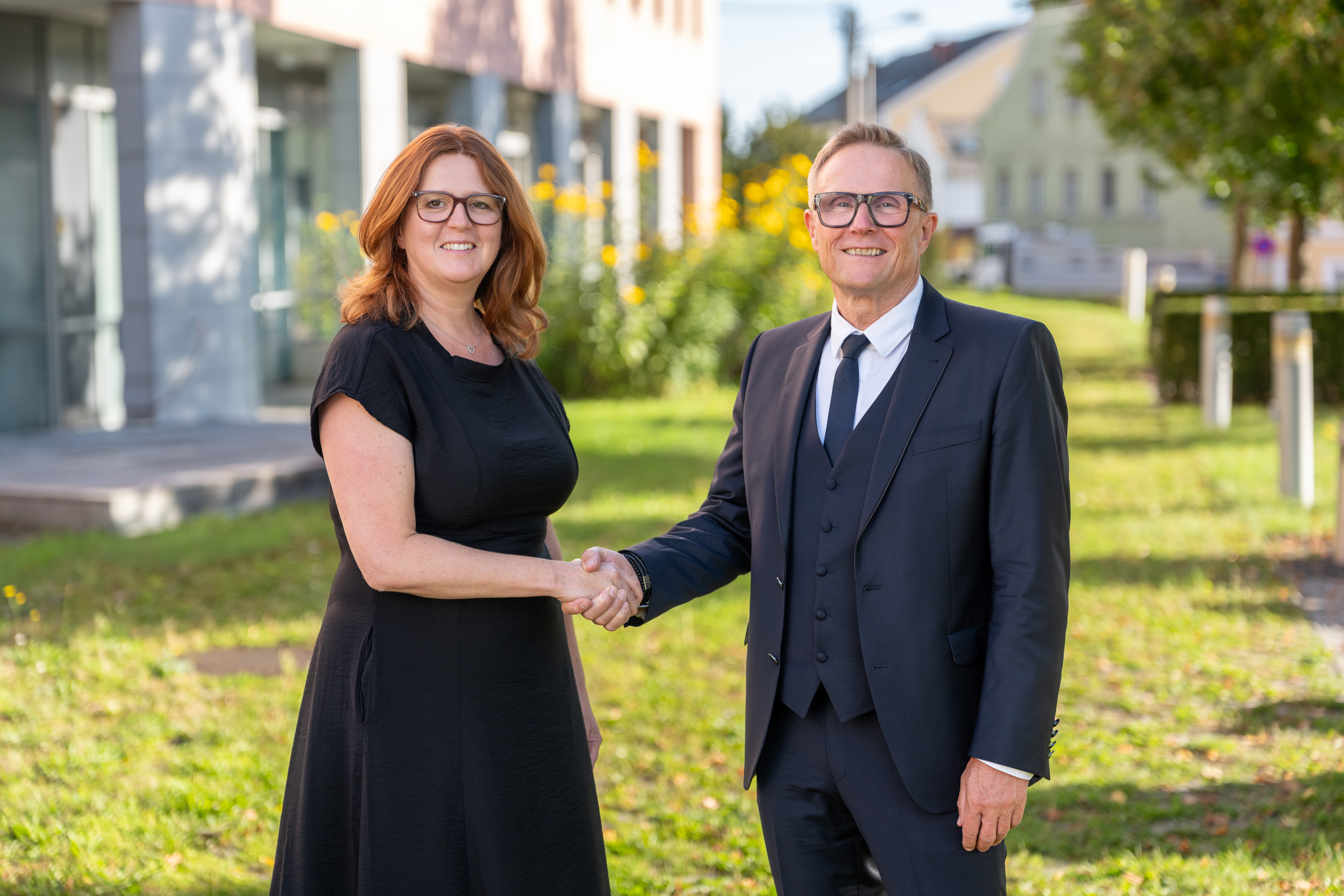 Frau Mag. Silvia S. und Herr Mag. Karl H. schütteln sich die Hände vor einem Gebäude in Österreich.